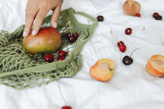 Hand arranging ripe mango and cherries in a green mesh bag on a white backdrop.