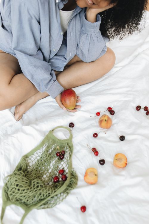 Free Woman in casual attire holding a mango with scattered fruits on white linen, evoking a relaxed summer vibe. Stock Photo