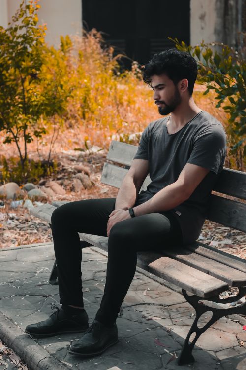Free Man in casual attire sitting pensively on a wooden bench in a park, surrounded by autumn foliage. Stock Photo