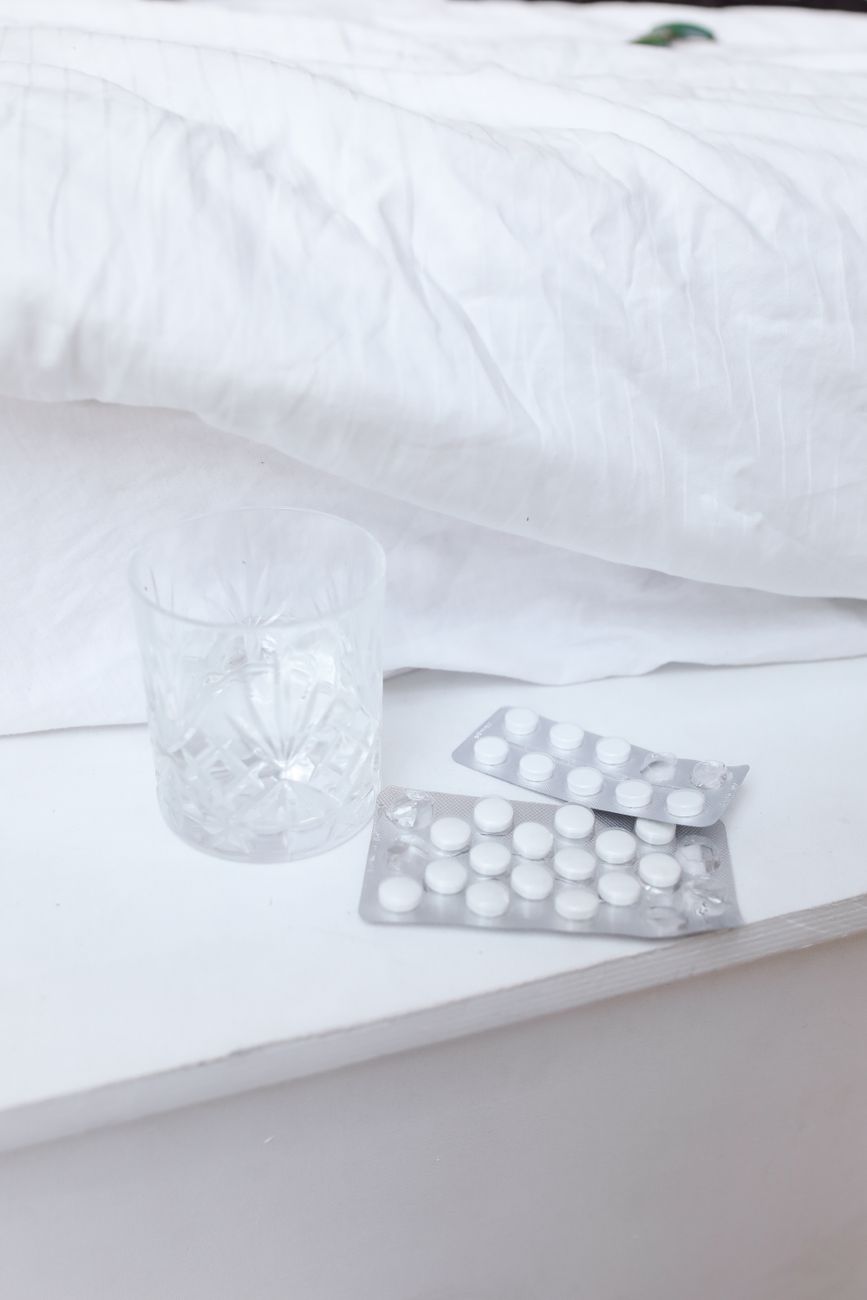 A clear glass and tablet blister packs on a white nightstand by a bed.