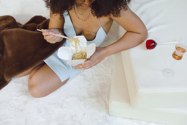 High Angle View Of A Woman Eating Noodles From A Paper Box On A Mattress