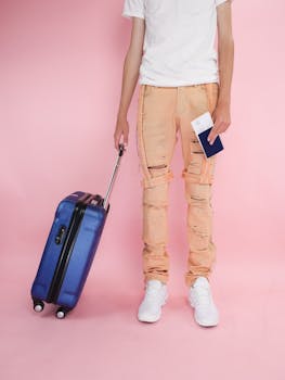 A trendy young adult holding a suitcase and passport, ready for travel, on a pink backdrop.