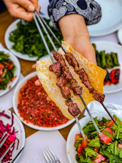 Free Hand holding grilled skewers with flatbread, surrounded by fresh salads and dips, showcasing vibrant Middle Eastern cuisine. Stock Photo