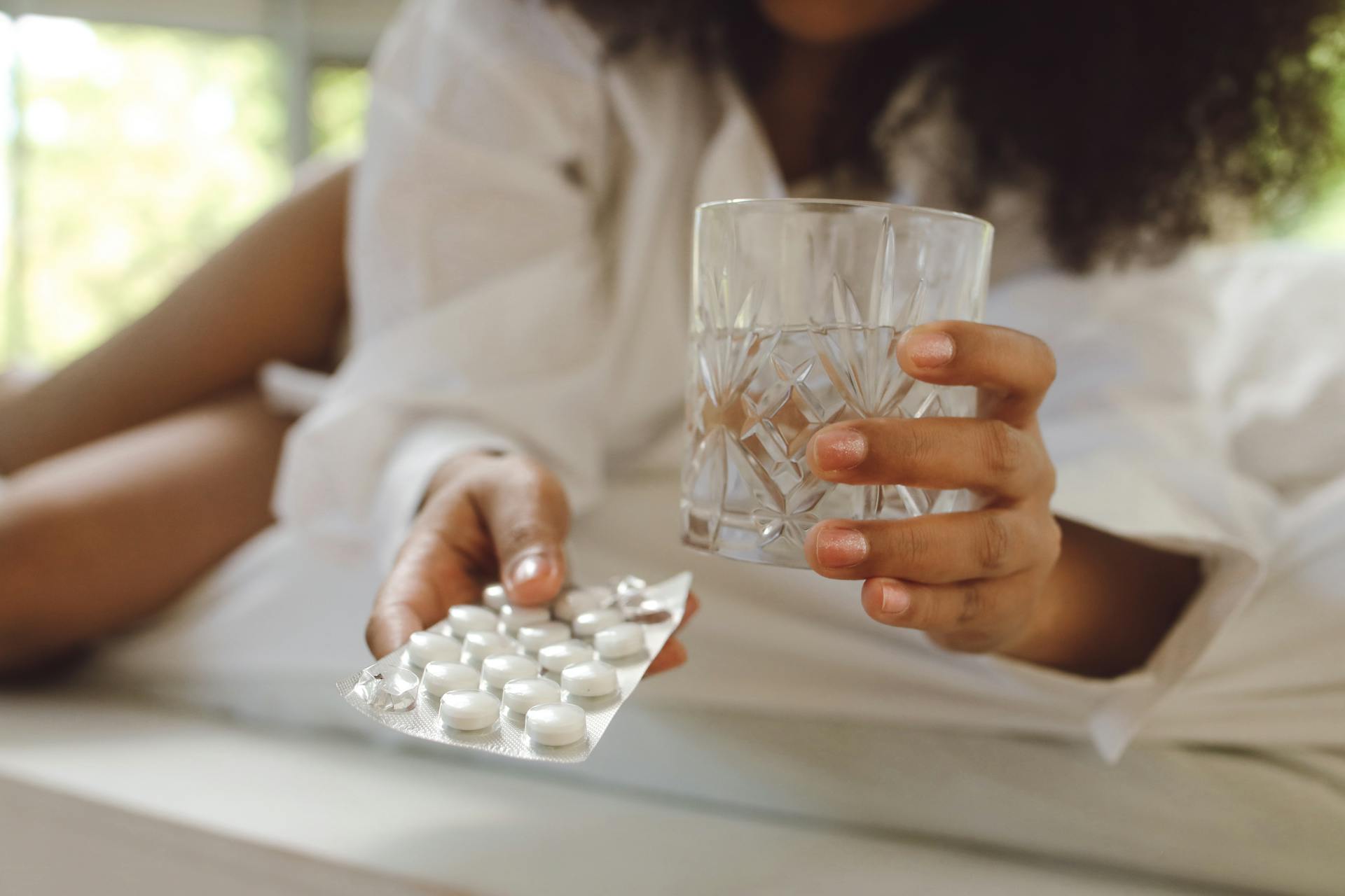 A woman in white long sleeves holds medication and a glass of water. Indoor lifestyle scene.