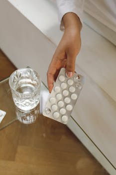 Close-up of a hand holding a blister pack with empty slots over a glass cup indoors.