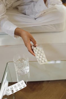 A person in white holds blister pack with a glass of water on table.