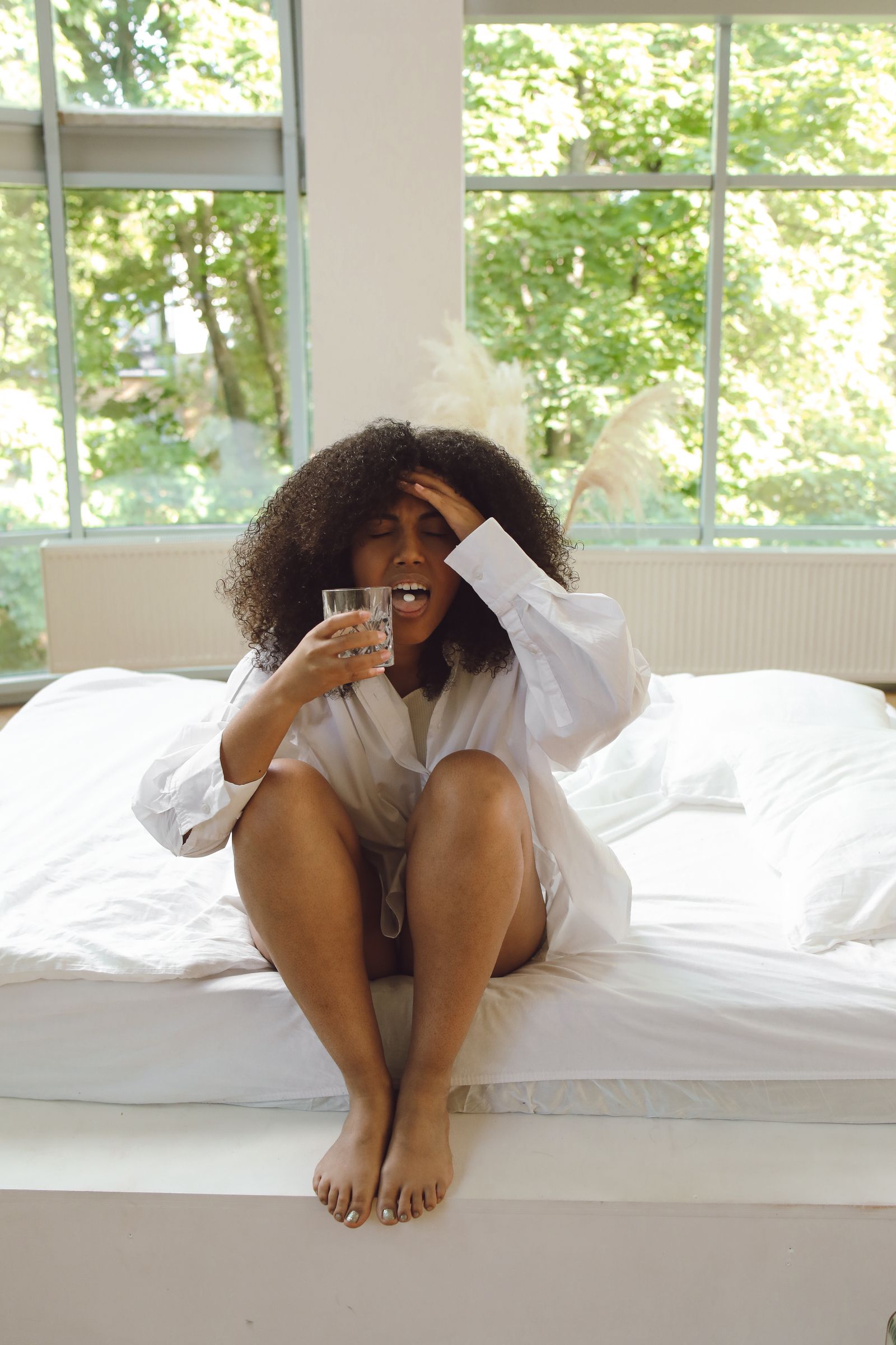 A Black woman sits on the edge of her bed in the morning light, one hand on her head and a glass of water in the other, looking exhausted after a poor night of sleep.