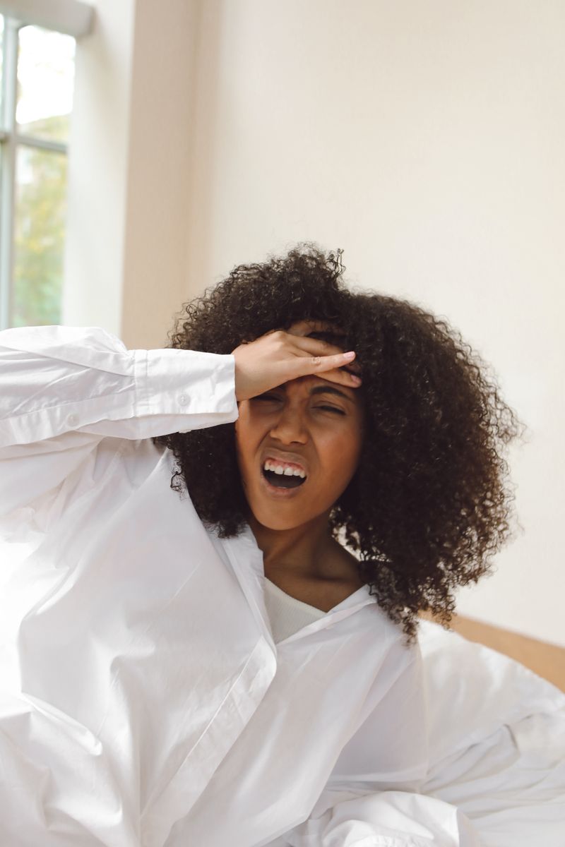 A Black woman in a white shirt holds the side of her head, eyes closed, in a sunlit room, a posture common during a vertigo or positional-dizziness episode.