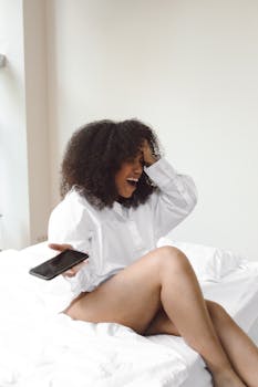 Joyful woman in white shirt laughs holding a smartphone on a bed.
