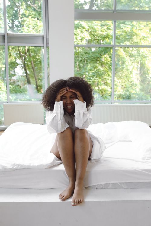 Free A woman in a white shirt sits on a bed, holding her head near large glass windows. Stock Photo