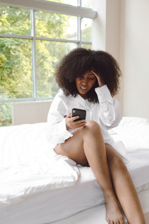 Free African American woman with curly hair using smartphone indoors, showing stress. Stock Photo