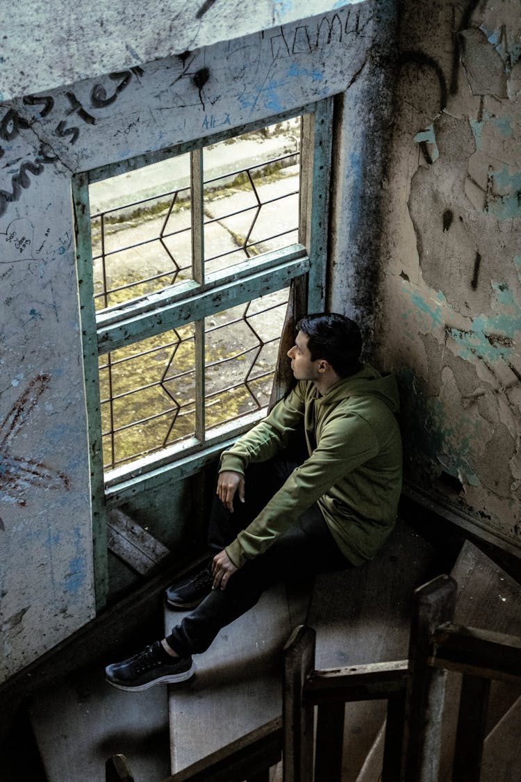 A Man Sitting On Stairs Inside An Abandoned House Looking Outside The Window