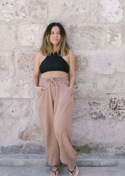 Woman in Black Top and Beige Pants with Hands in Pockets Leaning on Stone Wall