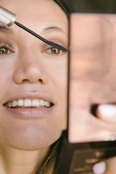 Close-up of a woman smiling as she applies mascara in front of a mirror.