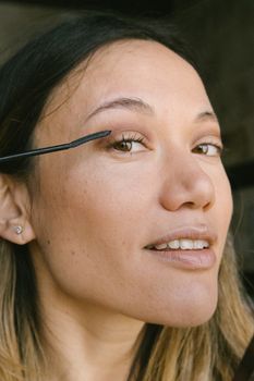 Close-up portrait of a woman applying mascara indoors. Neutral tones highlight her natural beauty.