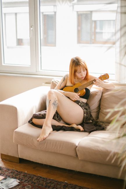 A young woman with tattoos plays the ukulele bare-footed on a cozy sofa by the window.