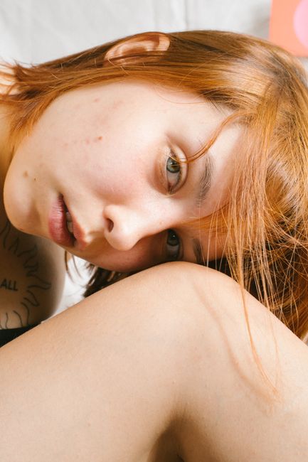 Close-up portrait of a young redhead woman resting her head on her knee, showcasing her tattoo and serene expression.