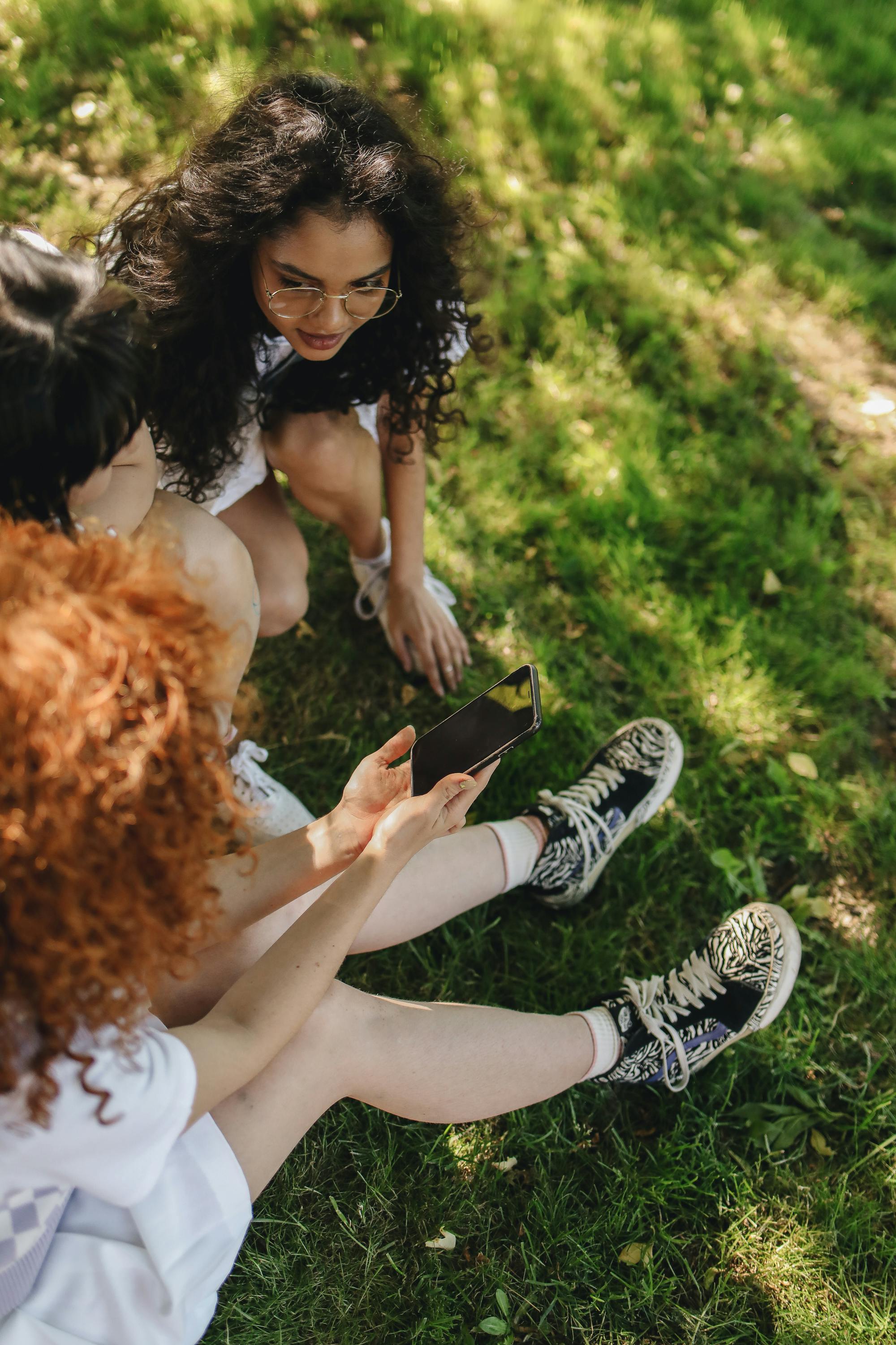 women sitting on a grassy ground while looking at the screen of a cellphone