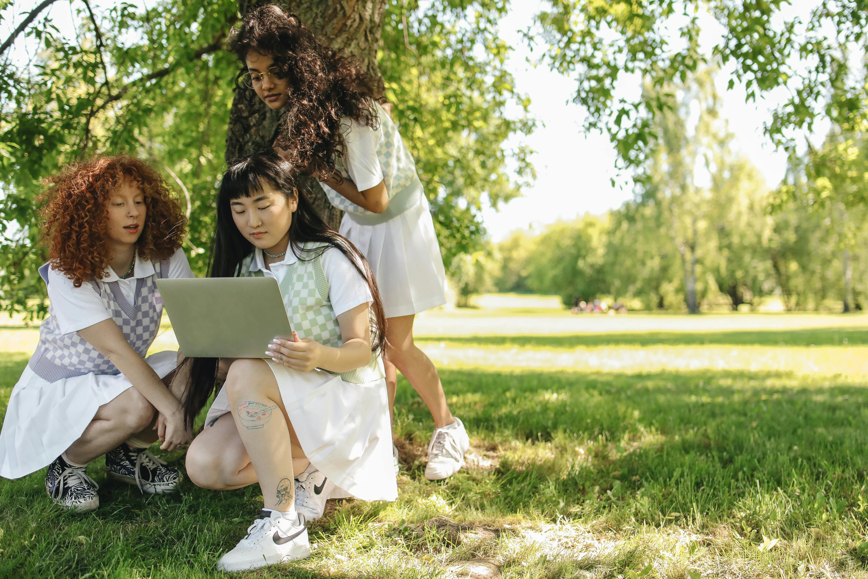 Three diverse young women in summer dresses interact with a laptop under a tree in a park.