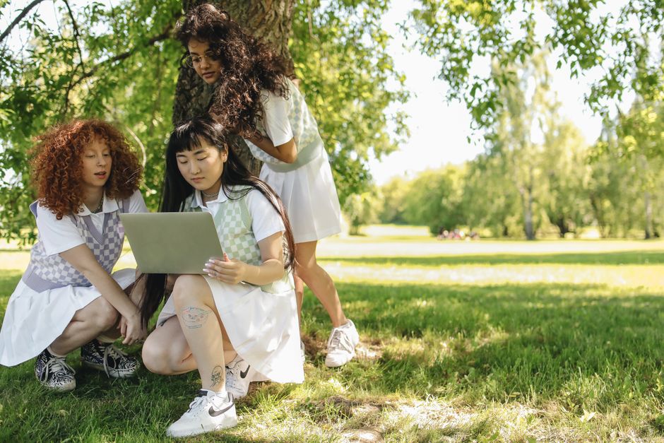 Perplexity AI vs ChatGPT: Research and Fact-Checking Workflow (2024) Three diverse young women in summer dresses interact with a laptop under a tree in a park.