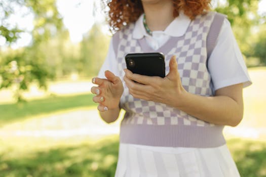 A woman holding a smartphone outdoors, wearing a sweater vest and polo shirt.
