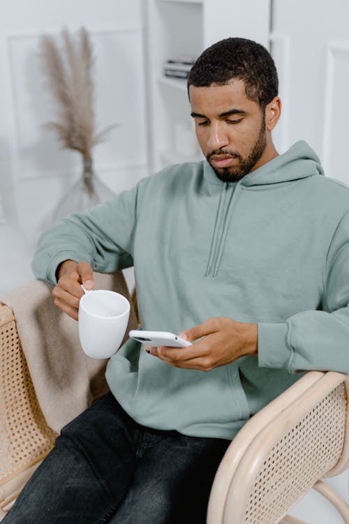 Free Man in Gray Hoodie Holding White Ceramic Mug and Looking at the Mobile Phone Stock Photo