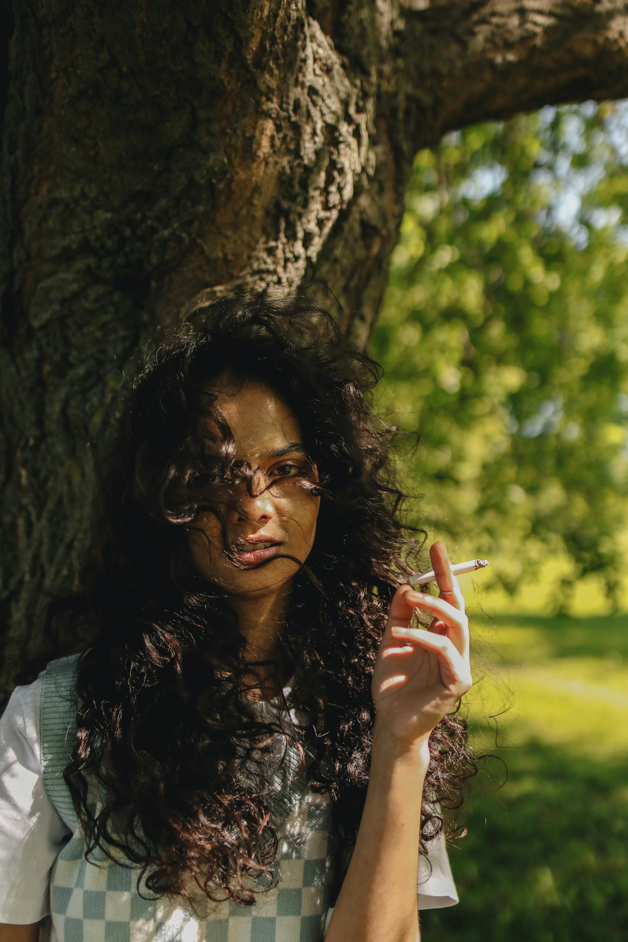 Portrait of a woman with curls smoking a cigarette under a tree on a sunny day.