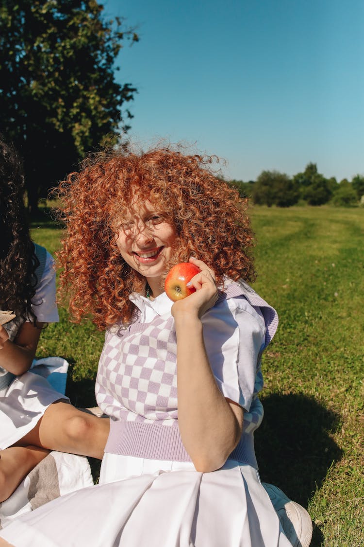 A Smiling Student Sitting On Grass Holding An Apple