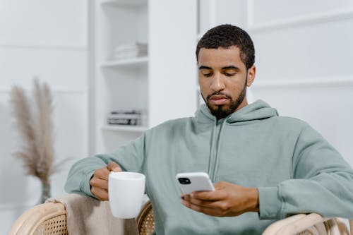 A Man in Gray Hoodie Holding a Cup of Coffee while Using a Smartphone