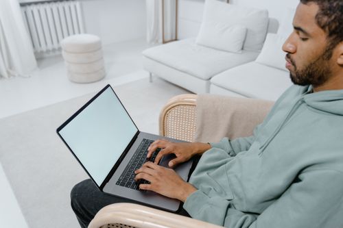 Free A man in a green hoodie sits and types on a laptop with a blank screen in a modern living room. Stock Photo