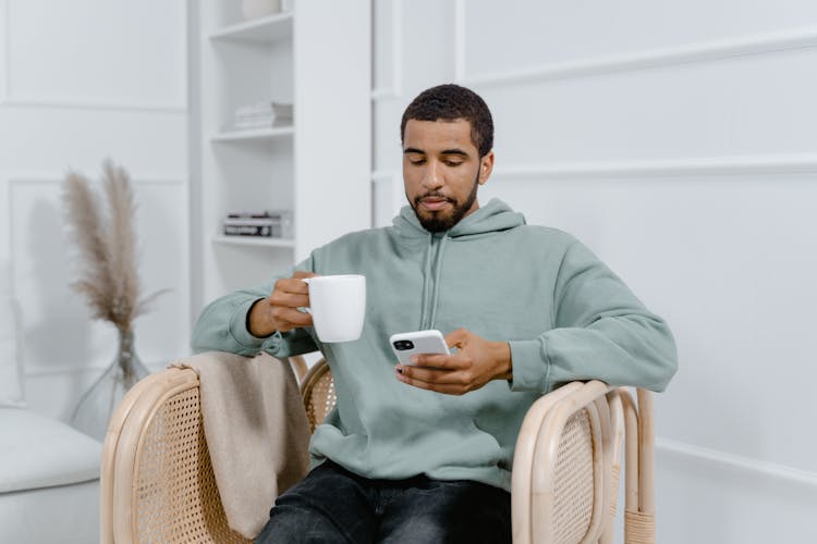 Man In A Hoodie Sitting On An Armchair Holding White Ceramic Mug