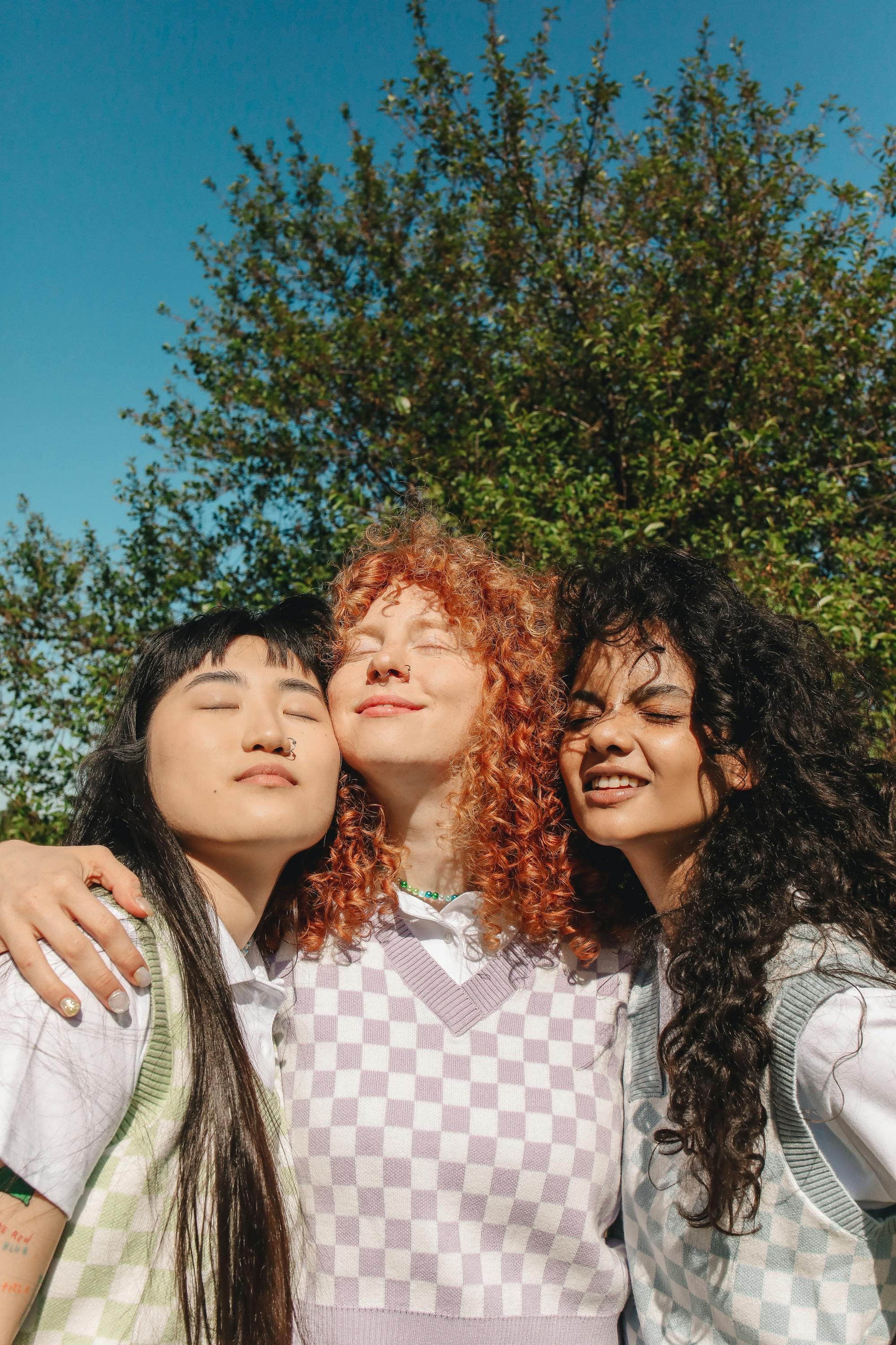 Three young women embracing outdoors, radiating friendship and happiness under a clear sky.