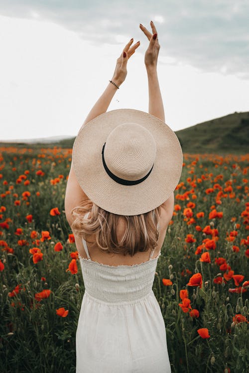 Back view of unrecognizable stylish female with blond hair in light clothes and hat standing with raised arms in field with blooming red poppy flowers and looking away in daytime