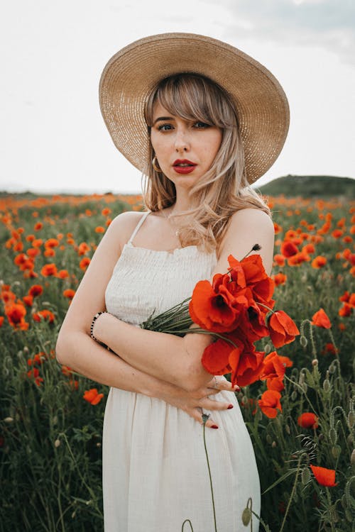 Thoughtful young female with red lips and blond hair in stylish clothes and hat standing with crossed arms and looking at camera against green field with red blossoming poppy flowers