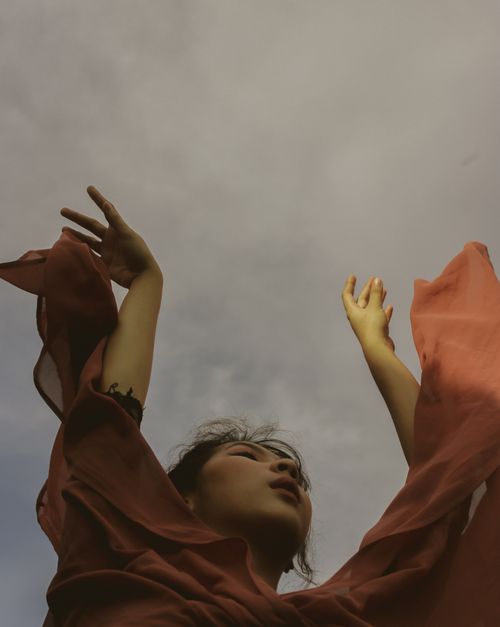 Free Woman with raised arms amidst flowing fabric, symbolizing freedom against a cloudy sky. Stock Photo