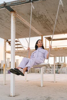 A woman in a pastel outfit sitting on a swing in a modern urban setting during sunset, exuding calm and tranquility.