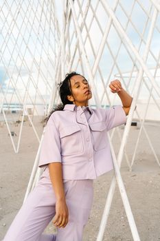 Woman in a lilac suit poses artistically against a geometric structure on a beach.