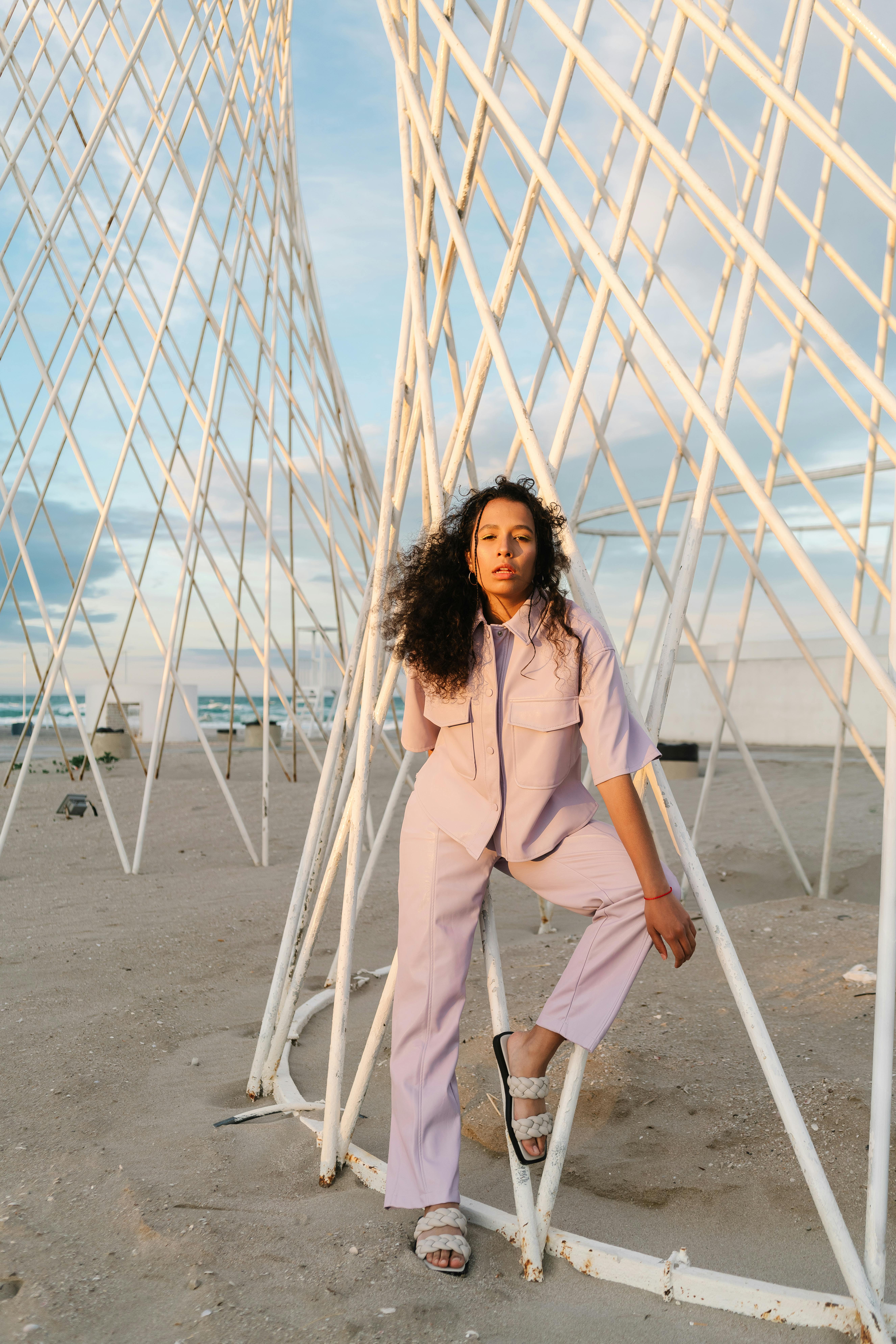 Stylish woman with afro hair posing confidently by artistic structures on a beach.