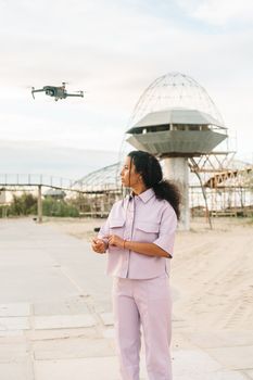 Stylish woman stands outdoors with drone, modern architecture in background.