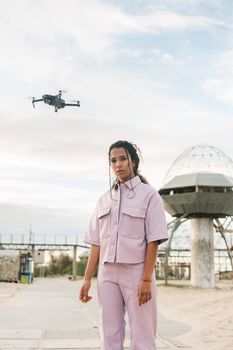 Young woman in pastel outfit stands with drone flying nearby in an urban scene.