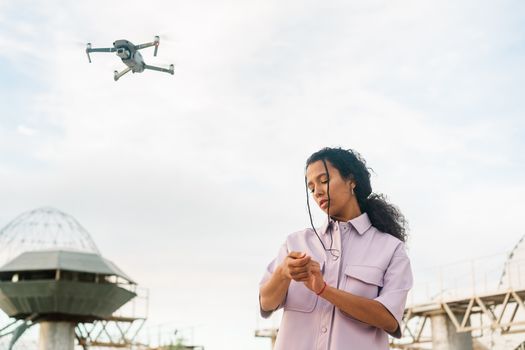 A woman controlling a drone in an open outdoor space with structures in the background.
