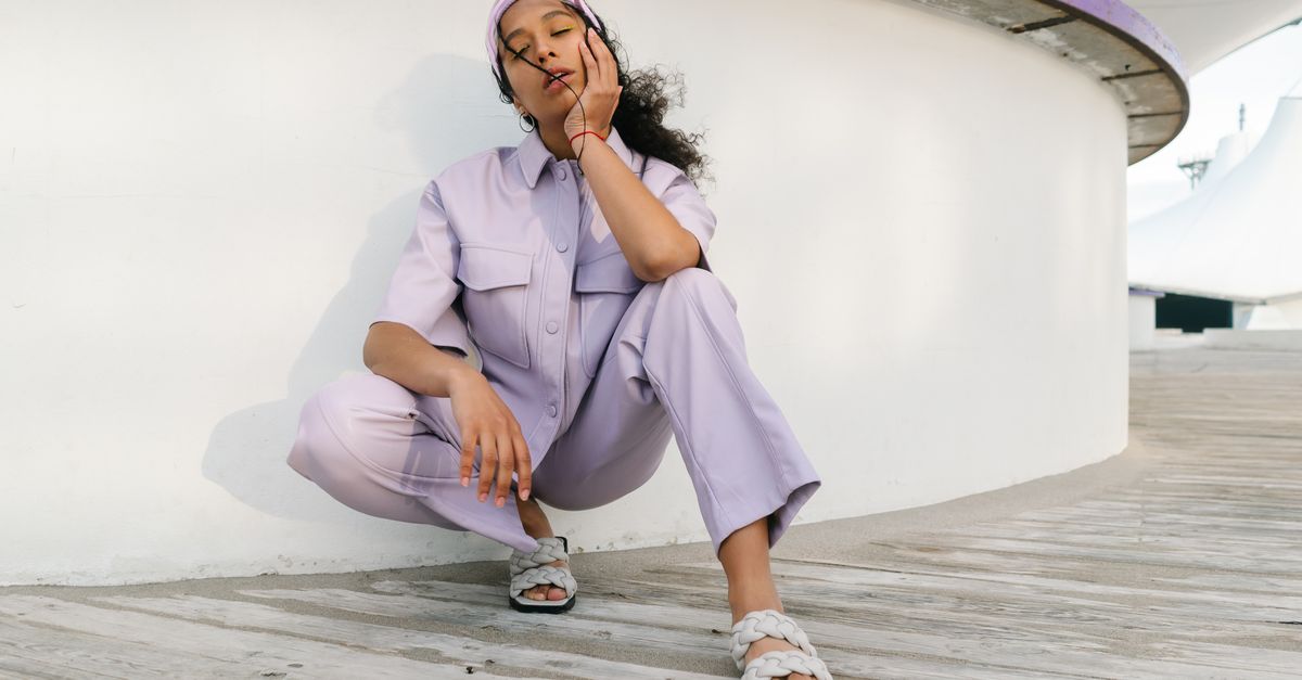 A fashionable woman in lavender attire posing outdoors with hand on cheek, showcasing modern style.