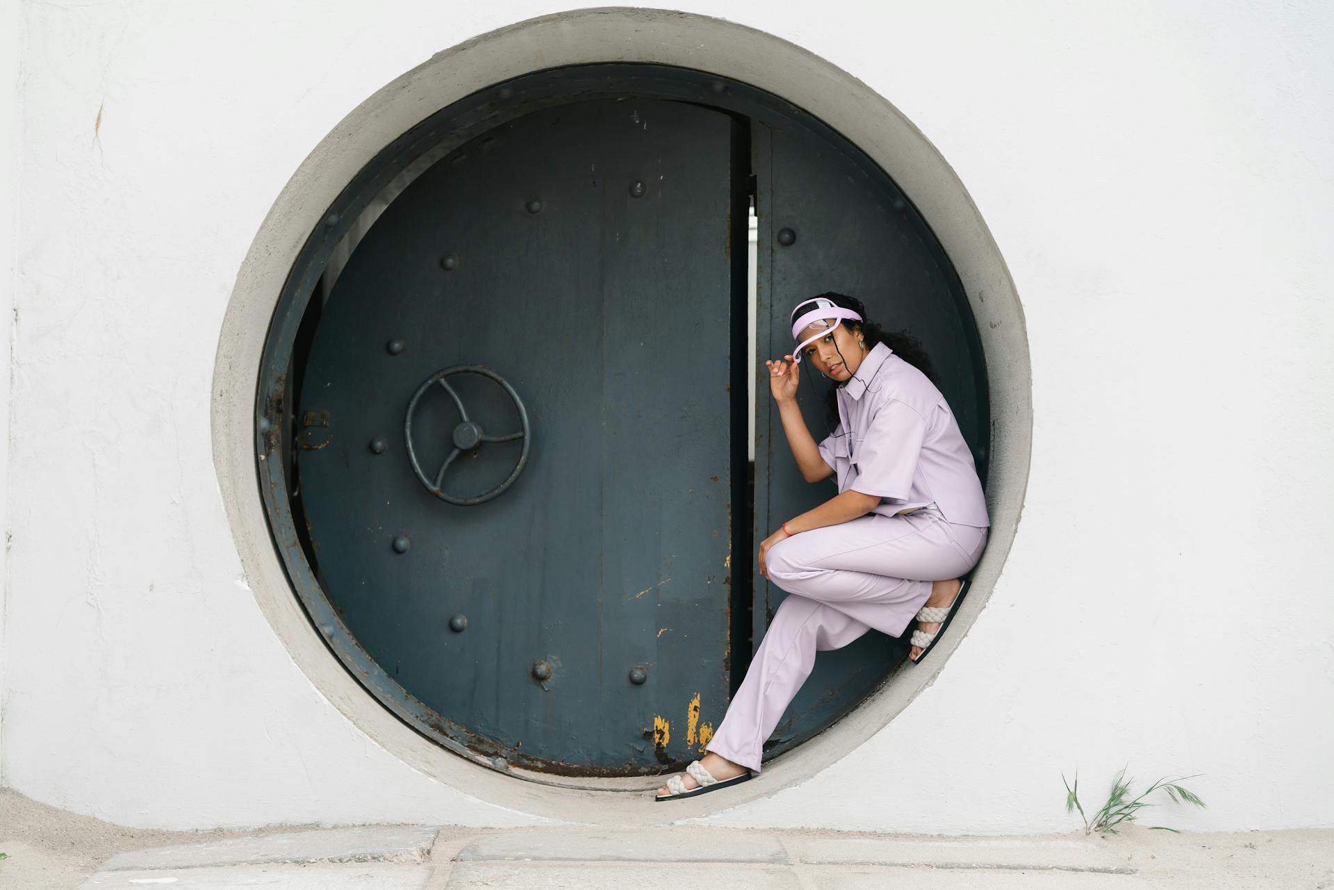 Stylish woman in pastel outfit poses confidently by a circular industrial vault door, adding a modern twist to an urban setting.