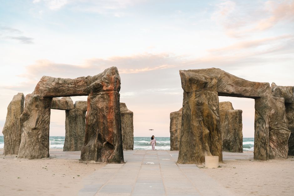 A Stonehenge-like structure on a beach with a person standing and the sea in the background at sunset.