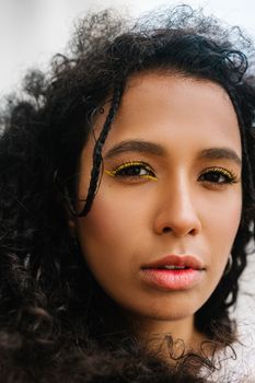 A close-up portrait of a woman with curly hair and bold yellow eye makeup, conveying confidence and style.