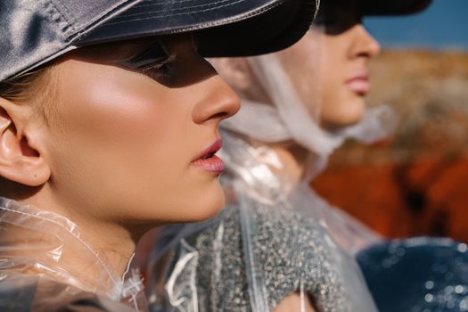 Profile view of two women wearing fashionable caps and shimmering makeup under sunlight.