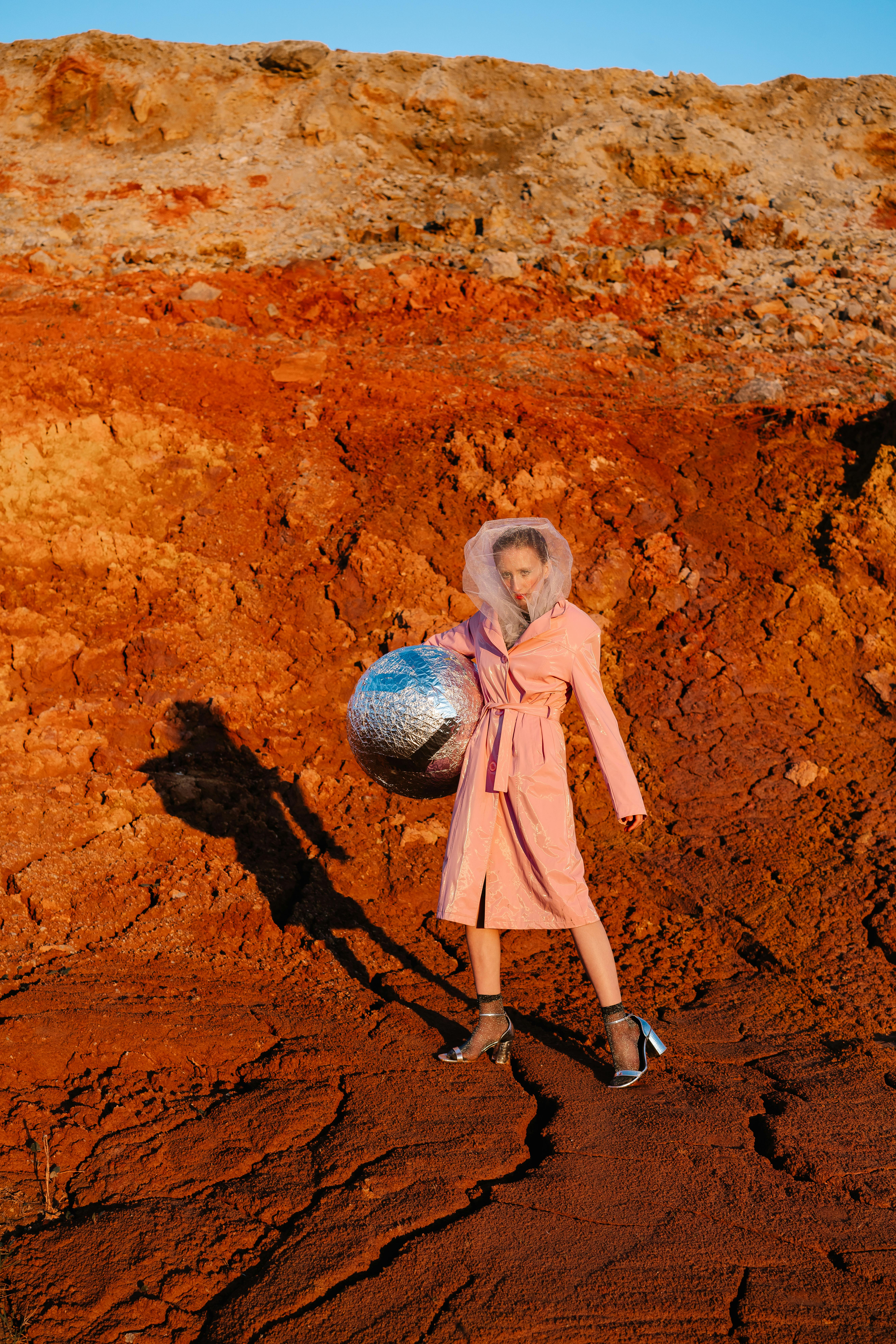 Stylish woman in pink coat holding mirrored ball in a rugged desert setting.
