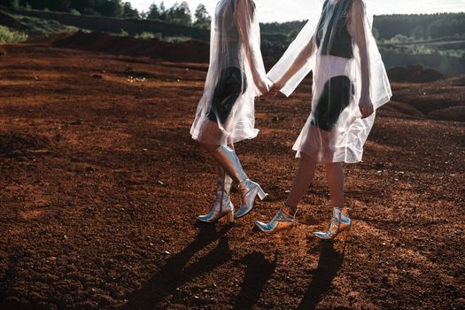 Two Women in Cellophane Coats and Silver Shoes Holding Hands and Walking on Red Sand
