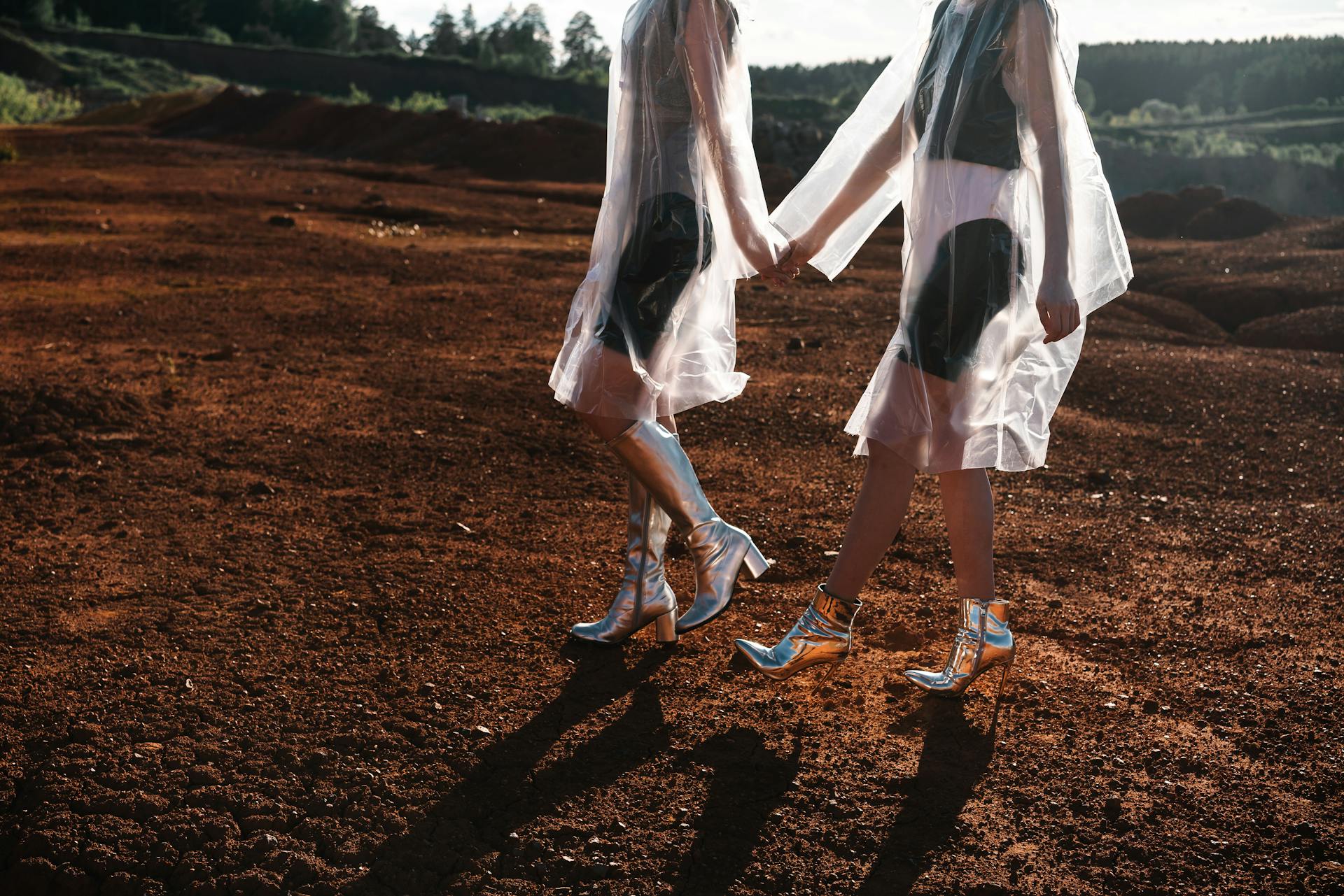 Two Women in Cellophane Coats and Silver Shoes Holding Hands and Walking on Red Sand