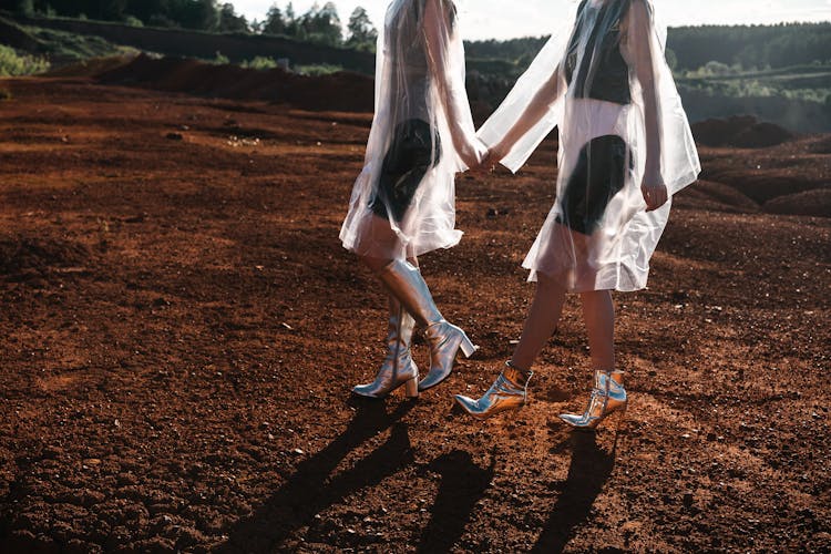 Two Women In Cellophane Coats And Silver Shoes Holding Hands And Walking On Red Sand